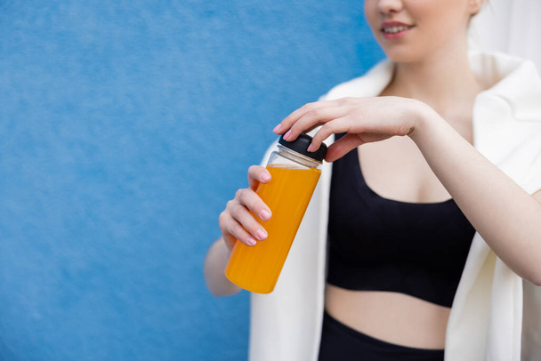 vista cortada de sportswoman sorrindo garrafa de abertura de suco de laranja perto da parede azul - Foto, Imagem