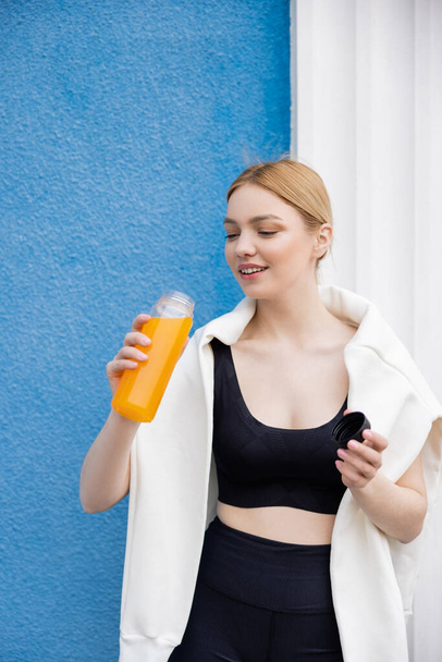 joven mujer deportiva sosteniendo botella con zumo de naranja fresco cerca de la pared azul - Foto, imagen