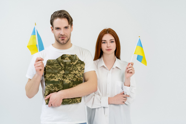 Young couple holding ukrainian flags and military uniform isolated on white  - 写真・画像