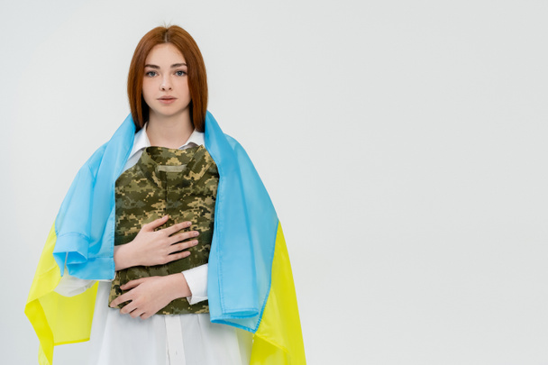 Κοκκινομάλλα γυναίκα με ουκρανική σημαία που κρατά στρατιωτική στολή απομονωμένη στα λευκά  - Φωτογραφία, εικόνα