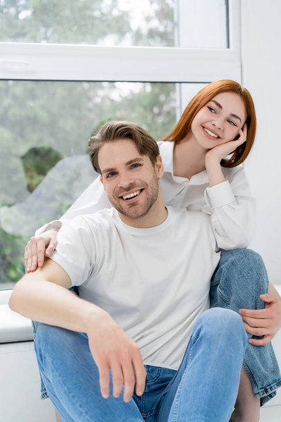 Smiling man looking at camera near red haired girlfriend at home  - Foto, Imagen