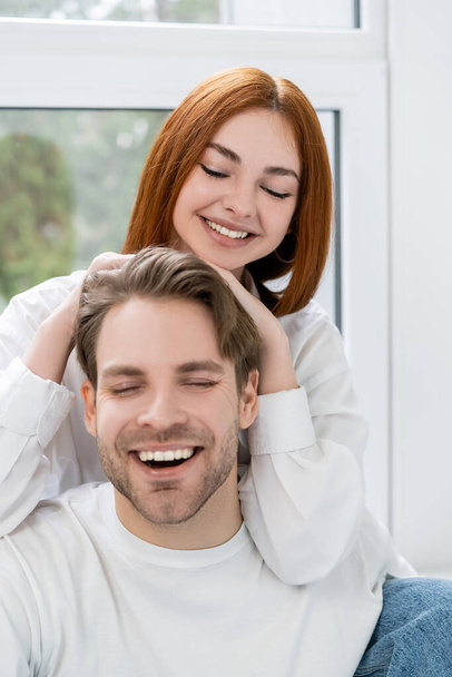 Positive redhead woman touching boyfriend at home  - 写真・画像