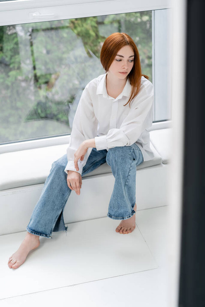 Pelirroja en jeans y camisa sentada en el alféizar de la ventana  - Foto, Imagen