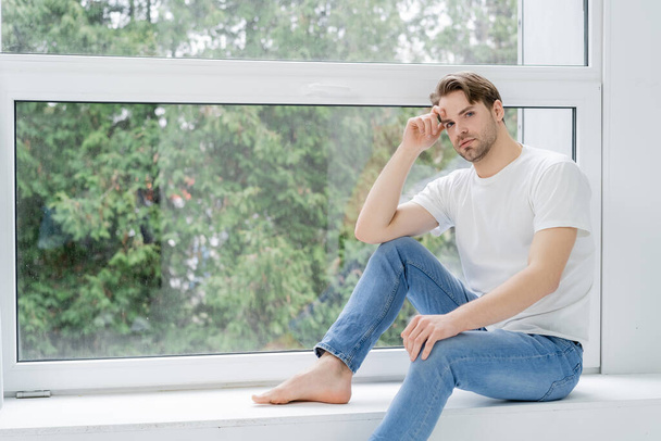 Barefoot man in jeans looking at camera near window at home  - Фото, зображення