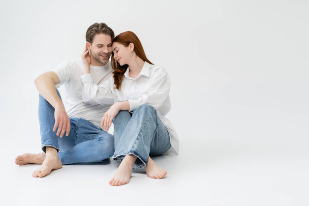 Barefoot woman touching ear of smiling boyfriend on white background - 写真・画像