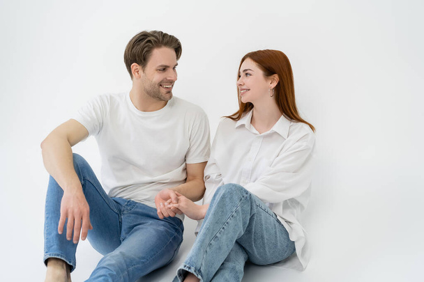 Positive couple in jeans holding hands while sitting on white background - Foto, imagen