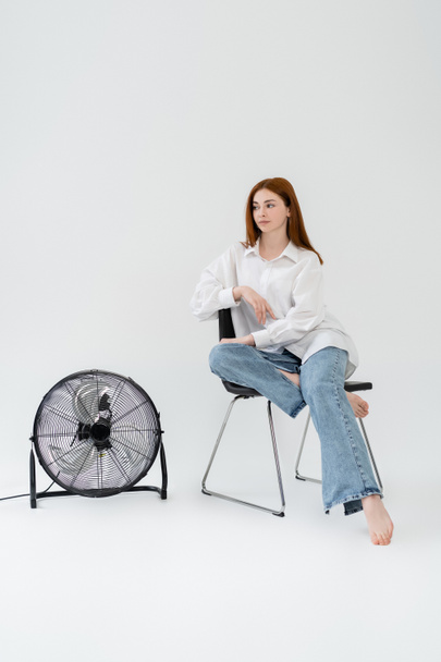 jonge roodharige vrouw zitten op stoel in de buurt elektrische ventilator op witte achtergrond - Foto, afbeelding