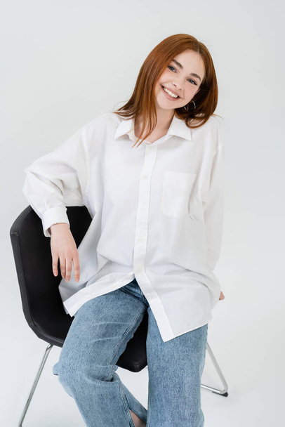 Cheerful young woman in shirt looking at camera while sitting on chair on white background - Foto, Imagen