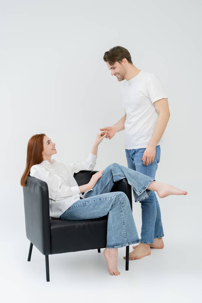 Side view of man holding hand of barefoot girlfriend sitting on armchair on white background - Fotografie, Obrázek