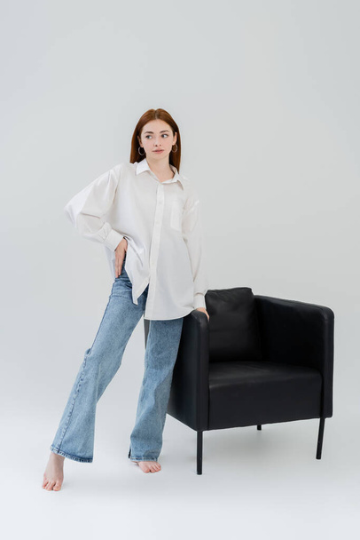 Full length of barefoot woman in jeans and shirt standing near armchair on white background - Photo, Image
