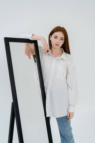 jonge vrouw in shirt posten in de buurt spiegel geïsoleerd op wit  - Foto, afbeelding