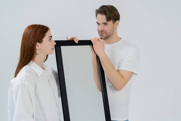 Joven pelirroja mirando novio cerca espejo aislado en blanco  - Foto, imagen