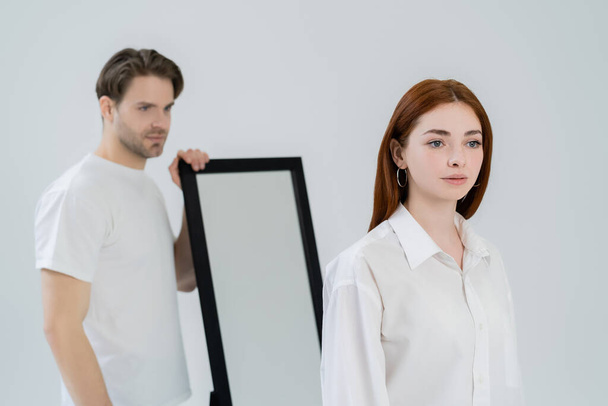 Young redhead woman standing near blurred boyfriend and mirror isolated on white  - Foto, imagen