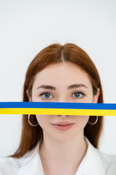 Redhead woman looking at camera near blue and yellow ribbon isolated on white  - Fotografie, Obrázek