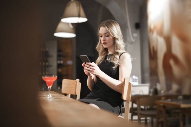 young woman drinks a cocktail in a bar and reads from her smartphone - Foto, Imagen