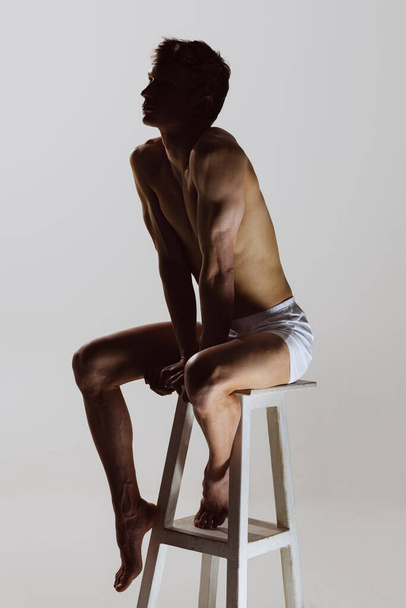 Looks sad, serious. One young shirtless sportive man in white boxer-briefs sitting on high chair isolated on grey background. Art, fashion, diversity, emotions - Photo, image