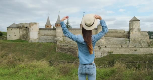 Mladá dívka stojí před starobylým hradem pevnosti, sundává si klobouk a rozpřahuje paže na stranu, objímajíc svět. Kamenets Podolsky, Ukrajina. Denní, zataženo, střední snímek. - Záběry, video