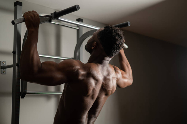 African american man with naked torso pulls up on horizontal bar in gym.  - 写真・画像