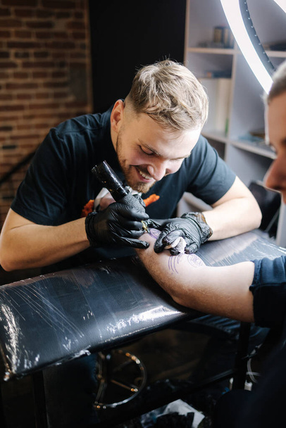 Ο δάσκαλος τατουάζ κάνει τατουάζ το χέρι ενός άντρα. Ασύρματη μηχανή τατουάζ, ασφάλεια και υγιεινή στο χώρο εργασίας. Κοντινό πλάνο της δουλειάς του καλλιτέχνη τατουάζ. Σαλόνι τατουάζ - Φωτογραφία, εικόνα