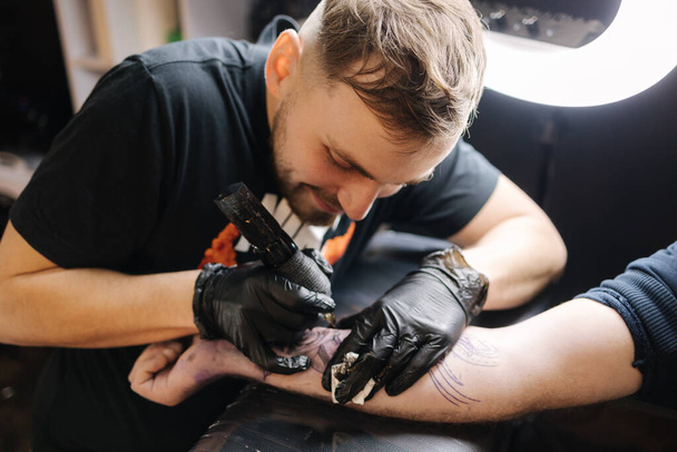 Mistrz tatuażu tatuuje męską dłoń. Bezprzewodowa maszyna do tatuażu, bezpieczeństwo i higiena pracy. Zbliżenie pracy tatuażysty. Salon tatuażu - Zdjęcie, obraz