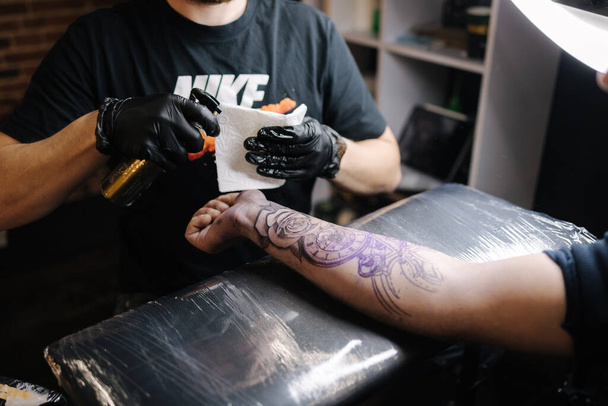 Close-up van een jonge professionele tattoo master introduceert zwarte inkt in de huid met behulp van een naald uit een tattoo machine. Tatoeage bij de hand - Foto, afbeelding
