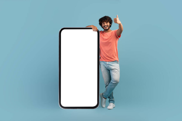 Positive indian guy posing by huge smartphone with blank screen - Fotó, kép