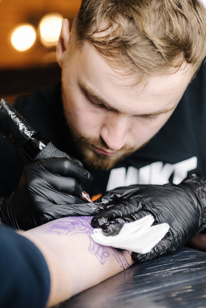 Κοντινό πλάνο του τατουατζή να κάνει μαύρο τατουάζ με μπογιά. Ο δάσκαλος δουλεύει με μαύρα αποστειρωμένα γάντια. Νεαρός καλλιτέχνης τατουάζ στην εργασία - Φωτογραφία, εικόνα