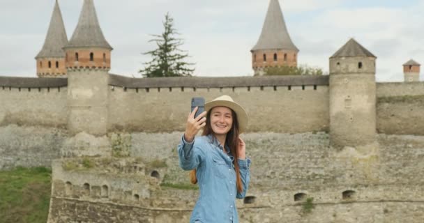 jong meisje met een hoed neemt een selfie met een telefoon tegen de achtergrond van een oud kasteel fort met torens. Medium shot, overdag, bewolkt, slow motion - Video