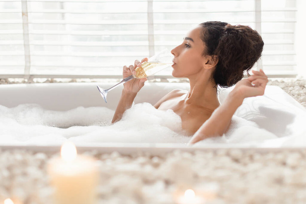 Sexy dama milenaria bebiendo champán mientras se encuentra en el baño caliente de burbujas en el hotel de lujo, espacio libre - Foto, Imagen