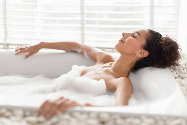 Vista lateral de una bonita mujer milenaria relajándose en un baño espumoso, tumbada en agua caliente burbujeante en casa, espacio para copiar - Foto, Imagen