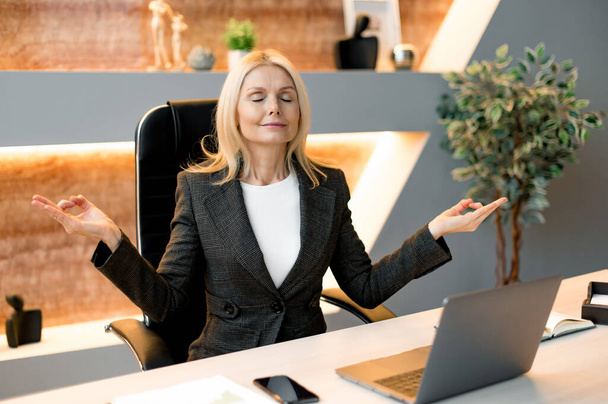 Middle aged positive calm relaxed caucasian blonde woman, boss, company ceo, secretary, working on laptop in modern office, resting and meditate while work, eyes closed, smiling, calmness concept - Photo, image