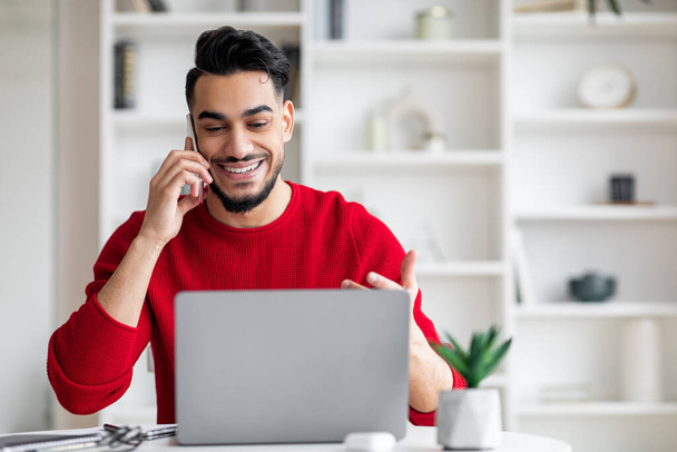 Uśmiechnięty, atrakcyjny arabski mężczyzna z brodą w czerwonym ubraniu rozmawia przez telefon i patrzy na laptopa. - Zdjęcie, obraz