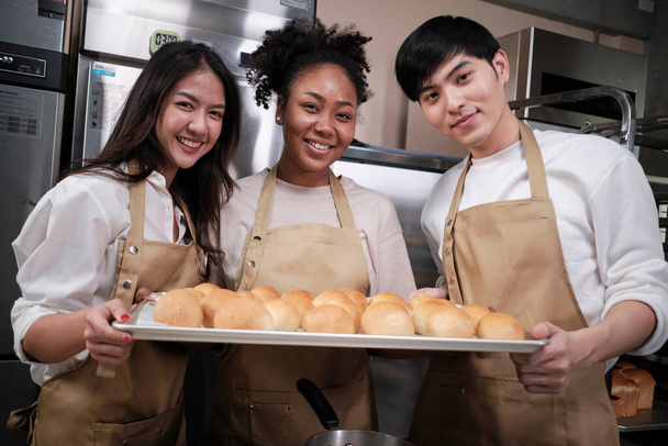Portret trzech początkujących partnerów cukierniczych patrzących w kamerę z wesołym uśmiechem i dumnych z tacy chleba w kuchni. Przyjaciel i założyciel piekarni żywności i świeżego codziennego piekarni małych firm. - Zdjęcie, obraz