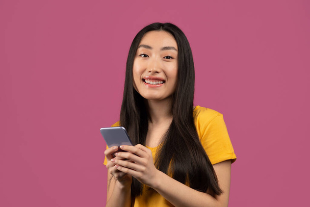Клевый гаджет и приложение. Счастливая корейская леди с помощью смартфона и улыбаясь в камеру, стоя на фоне фиолетовой студии - Фото, изображение