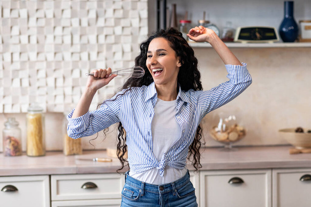 若いラテン語の女性が家で台所で料理をしながら、マイクとして泡立て器を保持し、歌を歌う - 写真・画像