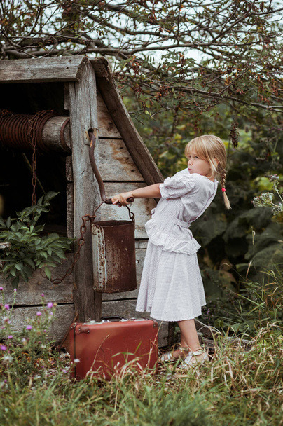 Beyaz benekli elbiseli neşeli küçük bir kızın portresi. Eski bir kuyudan su pompalamaya çalışıyor ve yaz mevsiminde kırsalda irkiliyor. Retro tarzı, çocukluk - Fotoğraf, Görsel