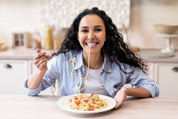 Happy excited latin woman eating tasty homemade pasta and smiling at camera, sitting at table in kitchen interior - Zdjęcie, obraz