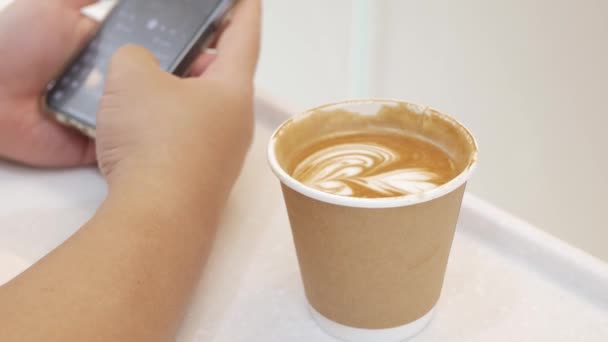 nach dem Handyspielen eine Tasse Kaffee auf weißem Tisch holen, Cappuccino heißen Kaffee in Pappbecher - Filmmaterial, Video