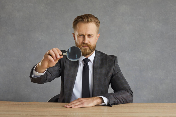 Serious focused businessman sitting at a table and looking through a magnifying glass. - Foto, Imagem