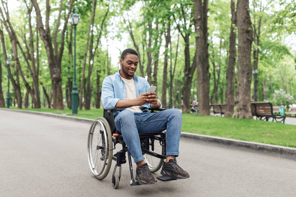 Comprimento total do jovem negro em cadeira de rodas usando telefone celular, verificando mensagens no parque, espaço livre - Foto, Imagem
