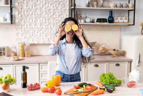 Playful woman having fun, putting orange slices to eyes at table with colorful vegetables in kitchen interior - Photo, Image