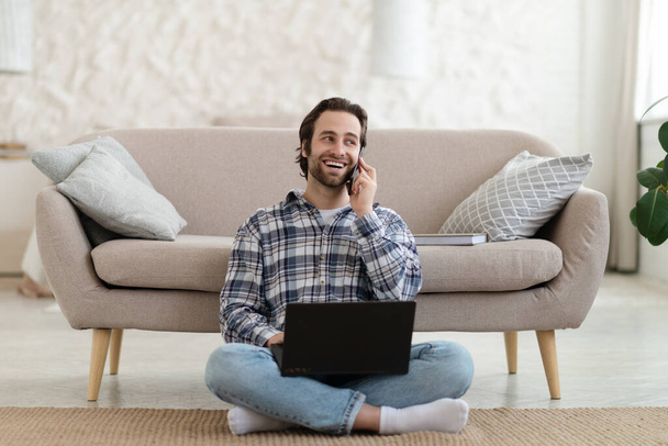 Happy millennial european guy with stubble in shirt calls by phone, works on laptop, sits on floor in living room - Foto, imagen