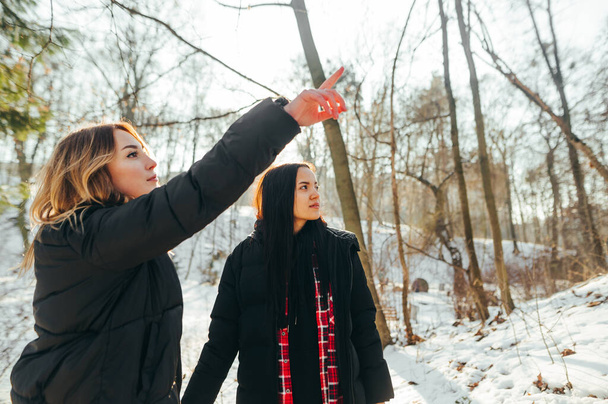 Two beautiful women in casual clothes stand in a snowy park during a walk, the girl shows her friend a finger to the side - Фото, изображение
