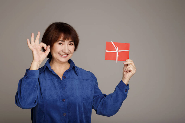 Happy middle aged mature woman in blue blouse holds red gift certificate isolated on grey background showing hand ok sign. People lifestyle holidays concept. - Foto, Bild