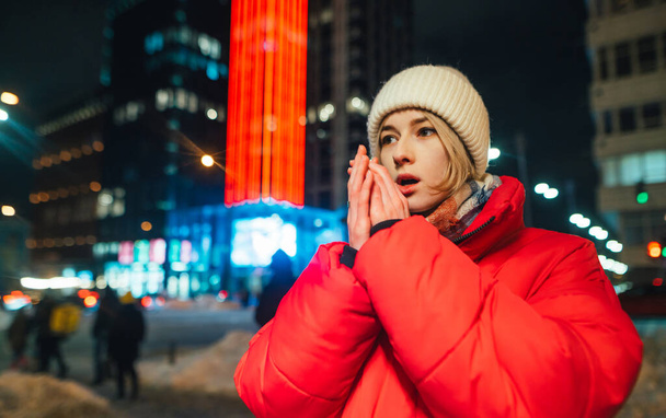Mooie vrouw verwarmt haar handen door erop te ademen buiten in sub-nul wintertemperaturen tegen de achtergrond van een nachtlandschap met moderne verlichte architectuur - Foto, afbeelding