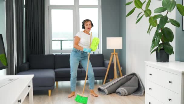 Een vrouw met een koptelefoon die het huis schoonmaakt en plezier heeft met dansen met een bezem en een washandje. Langzame beweging - Video