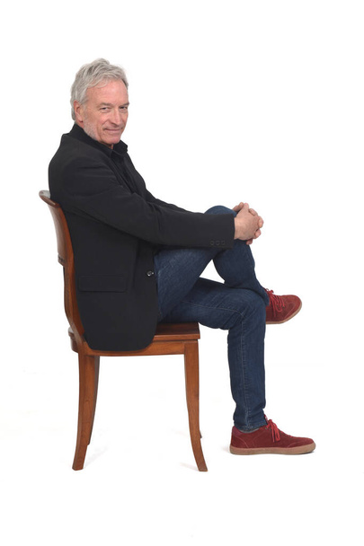 vista lateral de um homem sentado na cadeira com sapatilhas jeans e blazer, olhando para a câmera e pernas cruzadas no fundo branco - Foto, Imagem