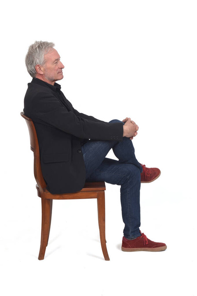 side view of a man sitting  on chair with sneakers jeans and blazer, legs crossed on white background - Foto, afbeelding