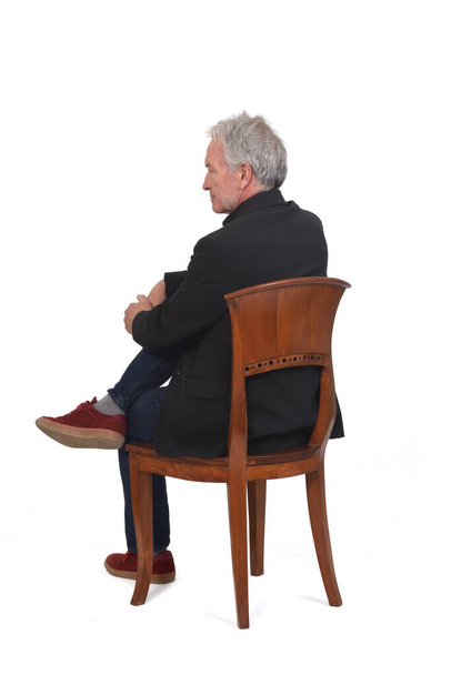 espalda y vista lateral de un hombre sentado en la silla con zapatillas vaqueros y chaqueta, piernas cruzadas sobre fondo blanco - Foto, Imagen