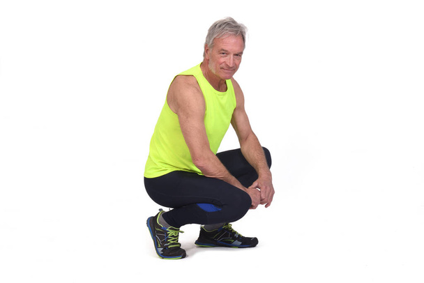 side view of a senior man with sportswear squatting looking at camera on white background - Foto, immagini